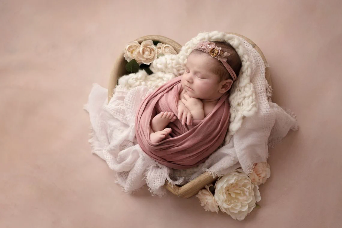 Sur un fond rose, une petite fille nouveau-né est dans un cœur en bois avec des fleurs autour. Elle porte un bandeau fleuri.