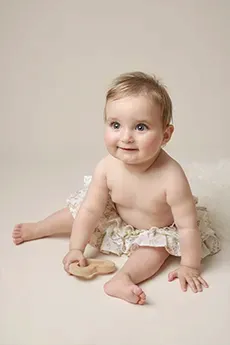 Bébé assis souriant en bloomer fleuri sur fond beige, tenant un coeur en bois, séance photo bébé à Dijon