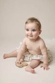 Bébé assis souriant en bloomer fleuri sur fond beige, tenant un jouet en bois, séance photo bébé à Dijon