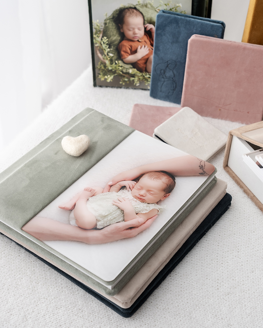 Supports d’images de votre photographe naissance à Dijon : albums photo en velours (vert sauge, rose, bleu, beige), avec une photo de naissance en couverture, d'autres supports en arrière-plan