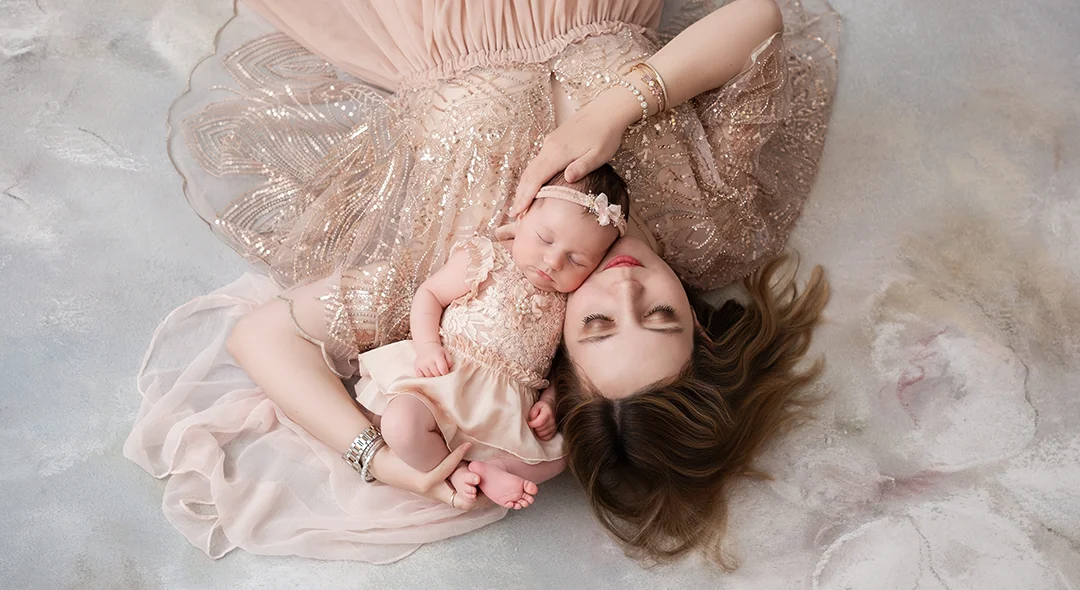 Photo d'une maman tenant son nouveau-né dans les mains. Elle et son bébé portent des robes délicates sur un fond fleuri tout doux
