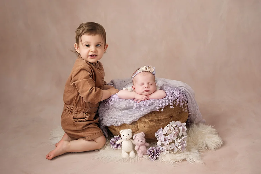 Photographe naissance à Dijon : nouveau-né endormi dans un panier en bois et dentelle lilas, avec sa grande soeur, sur un fond peint rose éclairé par une lumière douce