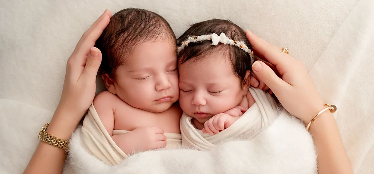 Jumeaux nouveau-nés endormis joue contre joue, entourés des mains protectrices de leur maman