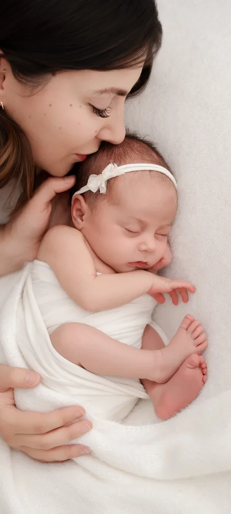 Photographe naissance à Dijon : maman déposant un baiser sur la tête de son nouveau-né endormi, emmailloté dans un lange blanc